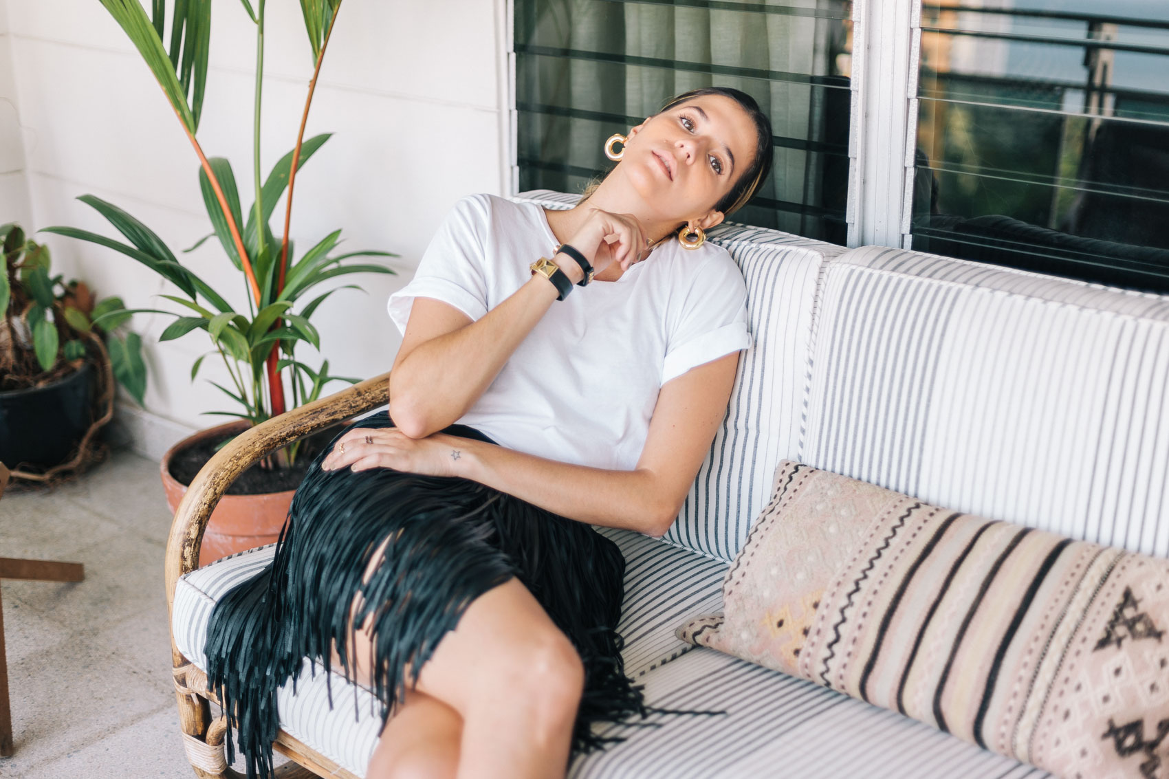 Maristella in her terrace patio, sitting on a rattan sofa, wearing a white tee from Cheap Monday with a leather mini skirt with fringe, leather bracelets with gold hardware from Yves Saint Laurent