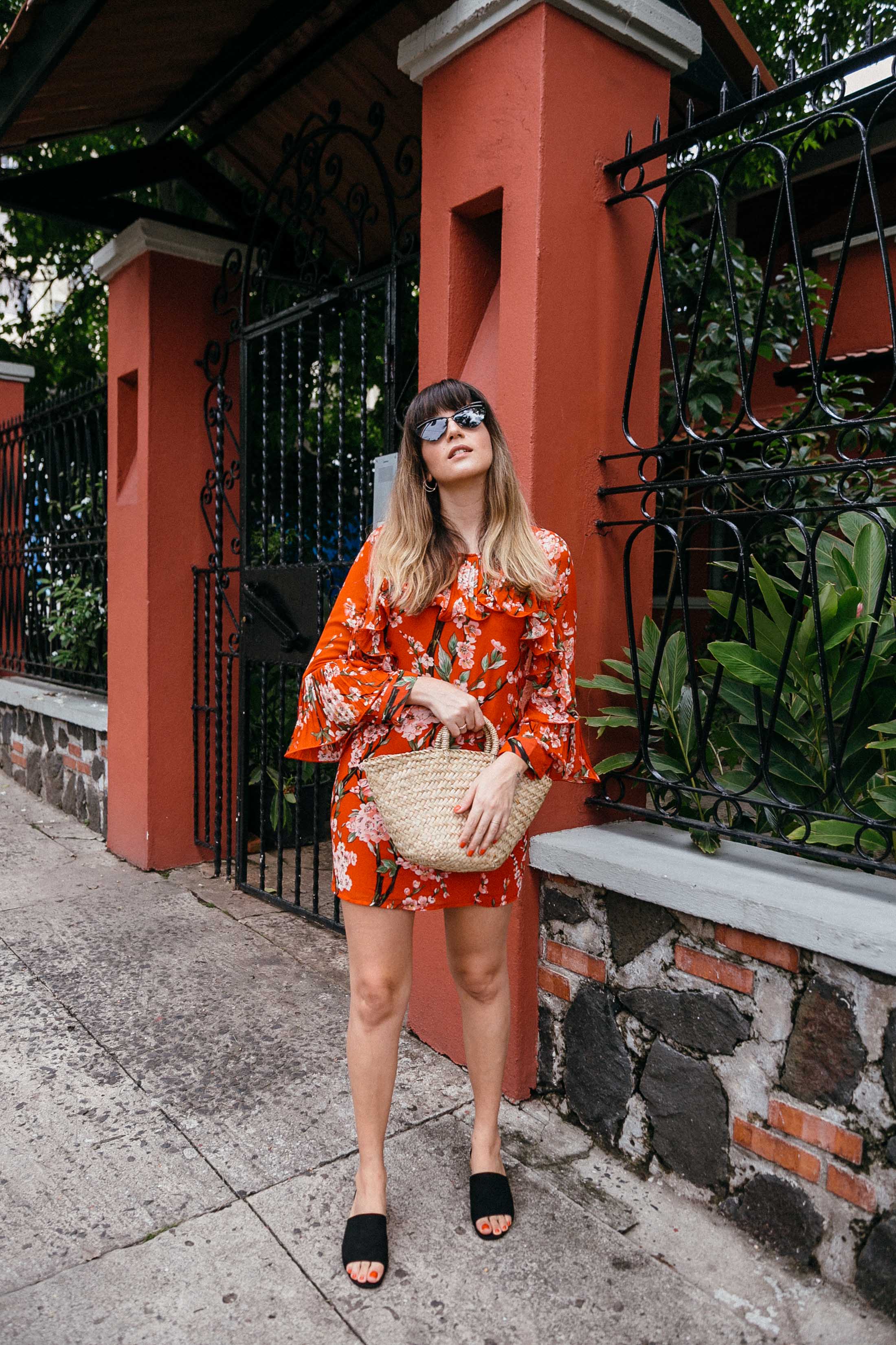 Maristella wears a red floral dress, black mules, straw bag and cat eye sunglasses