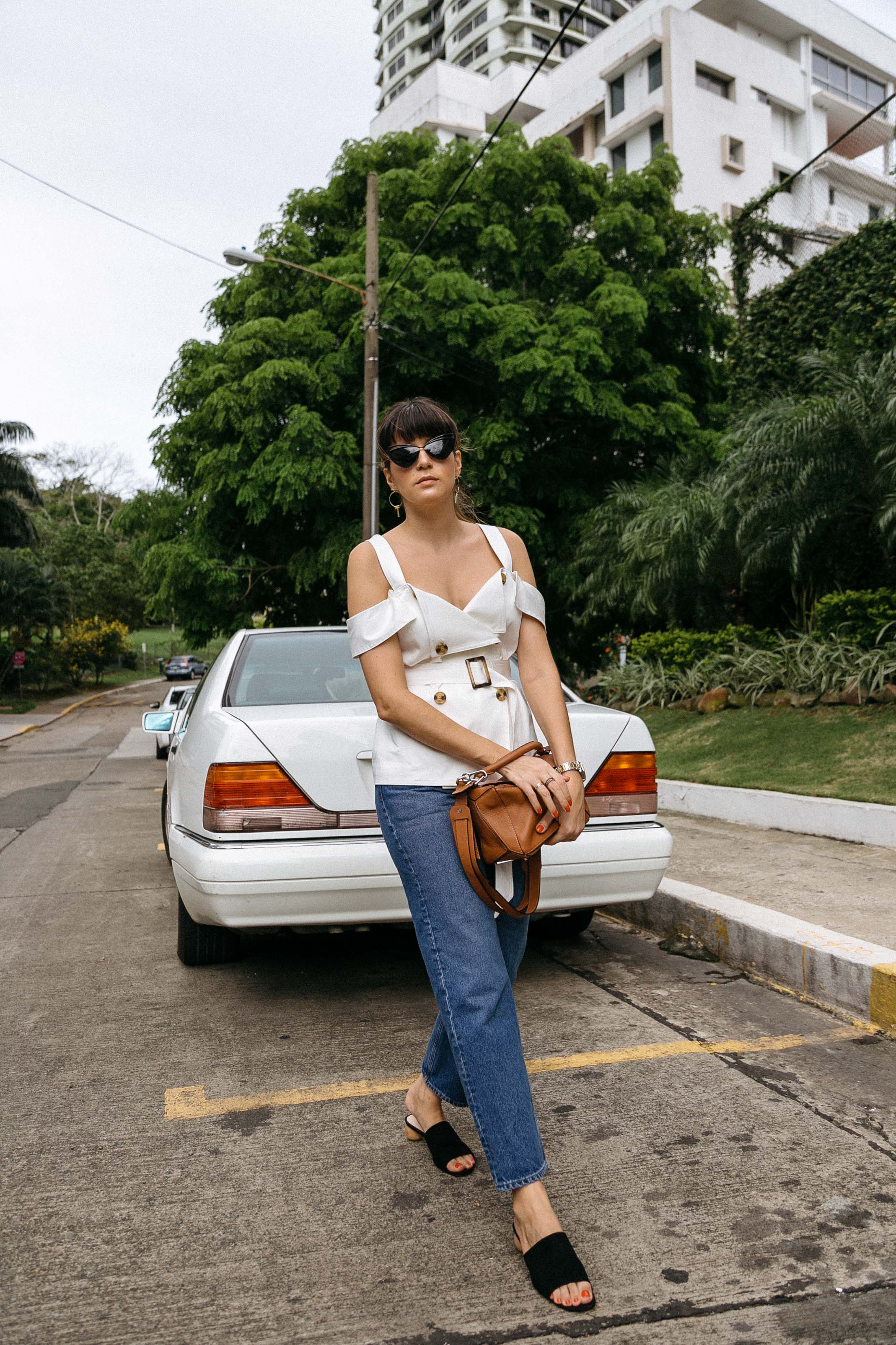 Maristella wears a white off the shoulders belted top with large buttons, straight leg jeans from COS, Mango wooden heel sandals and Loewe tan leather Puzzle bag